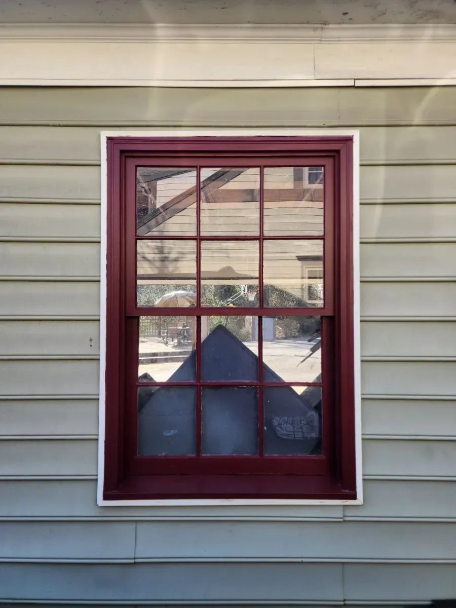 Red-framed window with white trim on a light green clapboard building, reflecting a dark shape and the outside.