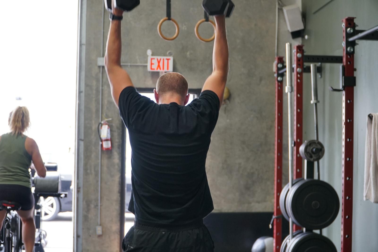 Man lifting dumbbells overhead in a gym, another person on a stationary bike in the background.