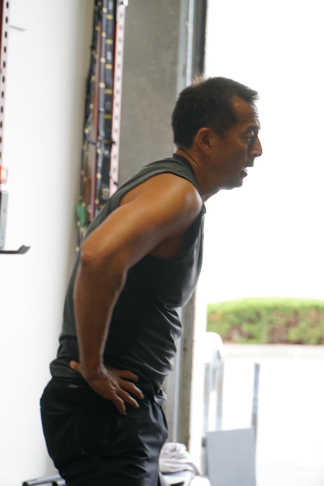 Man in workout clothes, hands on hips, leans forward, looking off to the side in a gym.