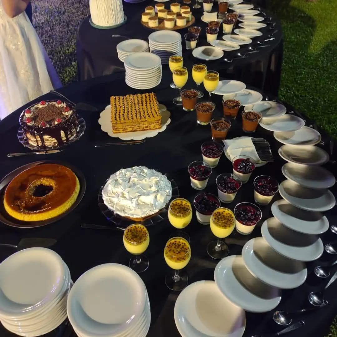Una mesa de postres tipo bufé con varios pasteles, flanes y vasitos dispuestos sobre manteles negros