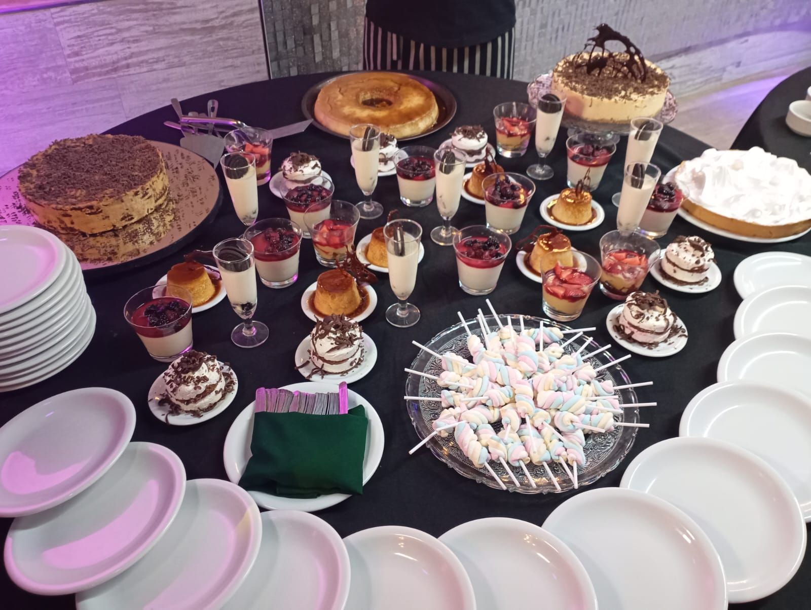 Una mesa de postres con pasteles, pudines en vasos, brochetas de malvaviscos y una pila de platos sobre un mantel negro.