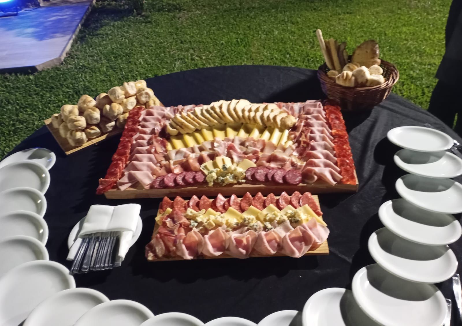 Dos tablas de embutidos, quesos y pan están dispuestas sobre un mantel negro al aire libre, con pilas de platos blancos.