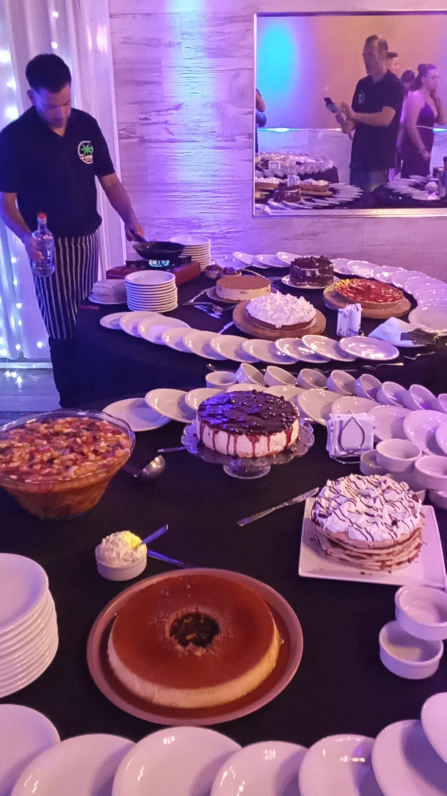 Un chef prepara la comida en una mesa de bufé de postres con pasteles, flan y platos apilados 