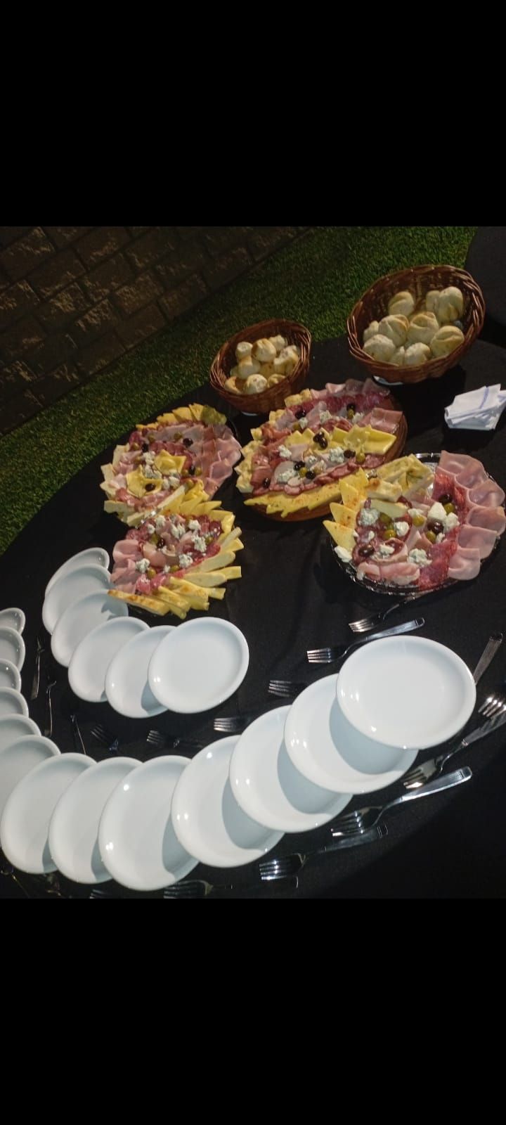 Un bufé con cuencos de embutidos, queso y pan sobre un mantel negro, junto a una pila de platos blancos.