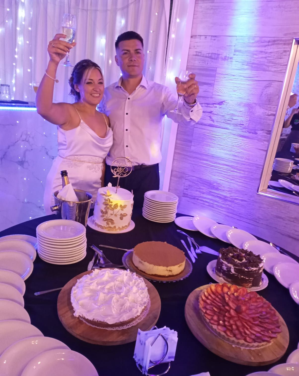 Una pareja sonriente brinda con champán detrás de una mesa llena de pasteles variados