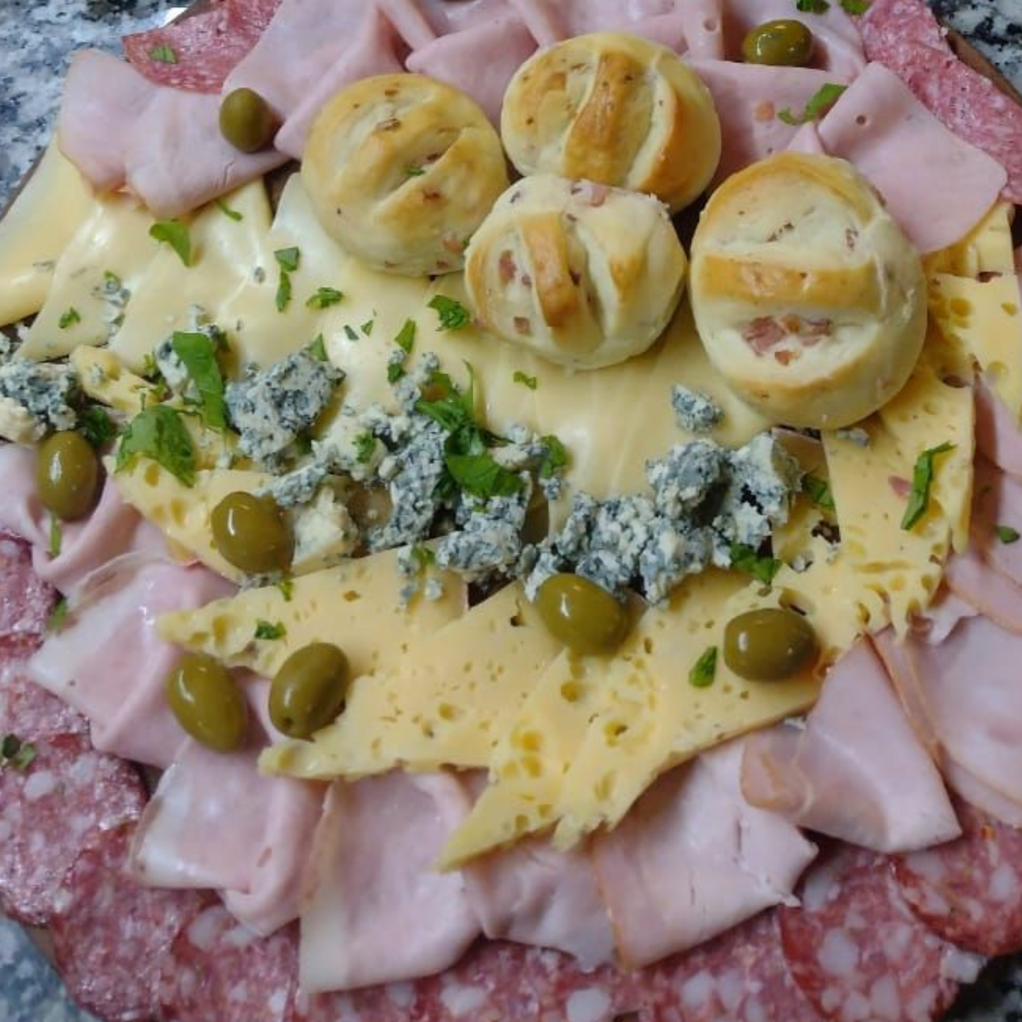 Vista aérea de una tabla de embutidos con carnes en lonchas, quesos variados, queso azul desmenuzado y aceitunas.