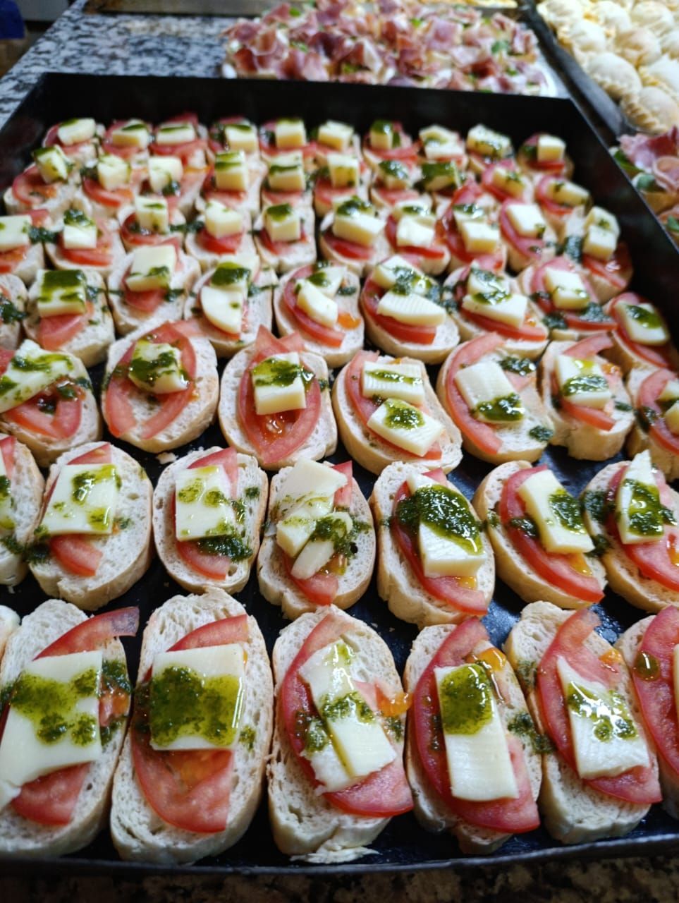 Una bandeja repleta de filas de bruschetta caprese cubiertas con rodajas de tomate, queso mozzarella y pesto verde.
