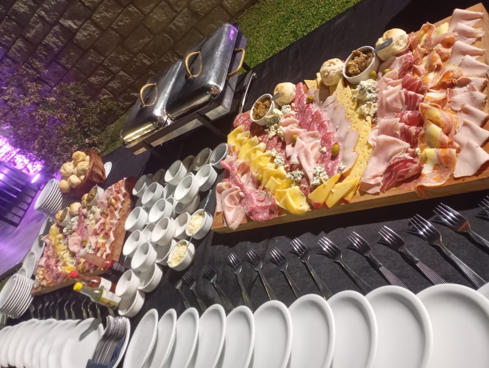 Una gran mesa de catering con varias tablas de madera con embutidos, carnes, quesos y pilas de platos sobre un mantel negro.