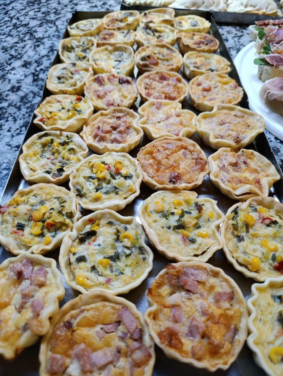 Una bandeja repleta de pequeñas quiches horneadas, algunas con maíz y verduras, otras con trozos de jamón a la vista.