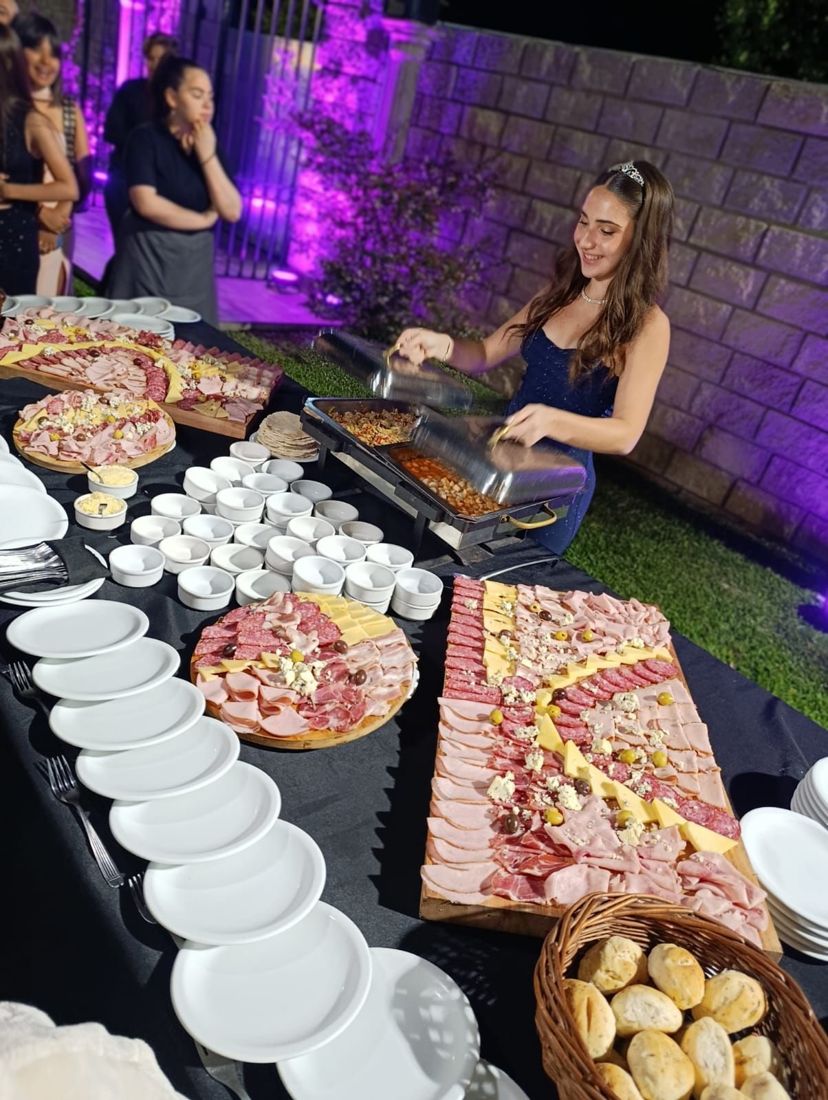 Un camarero prepara comida caliente en un bufé al aire libre que incluye grandes bandejas de embutidos, queso y pan.