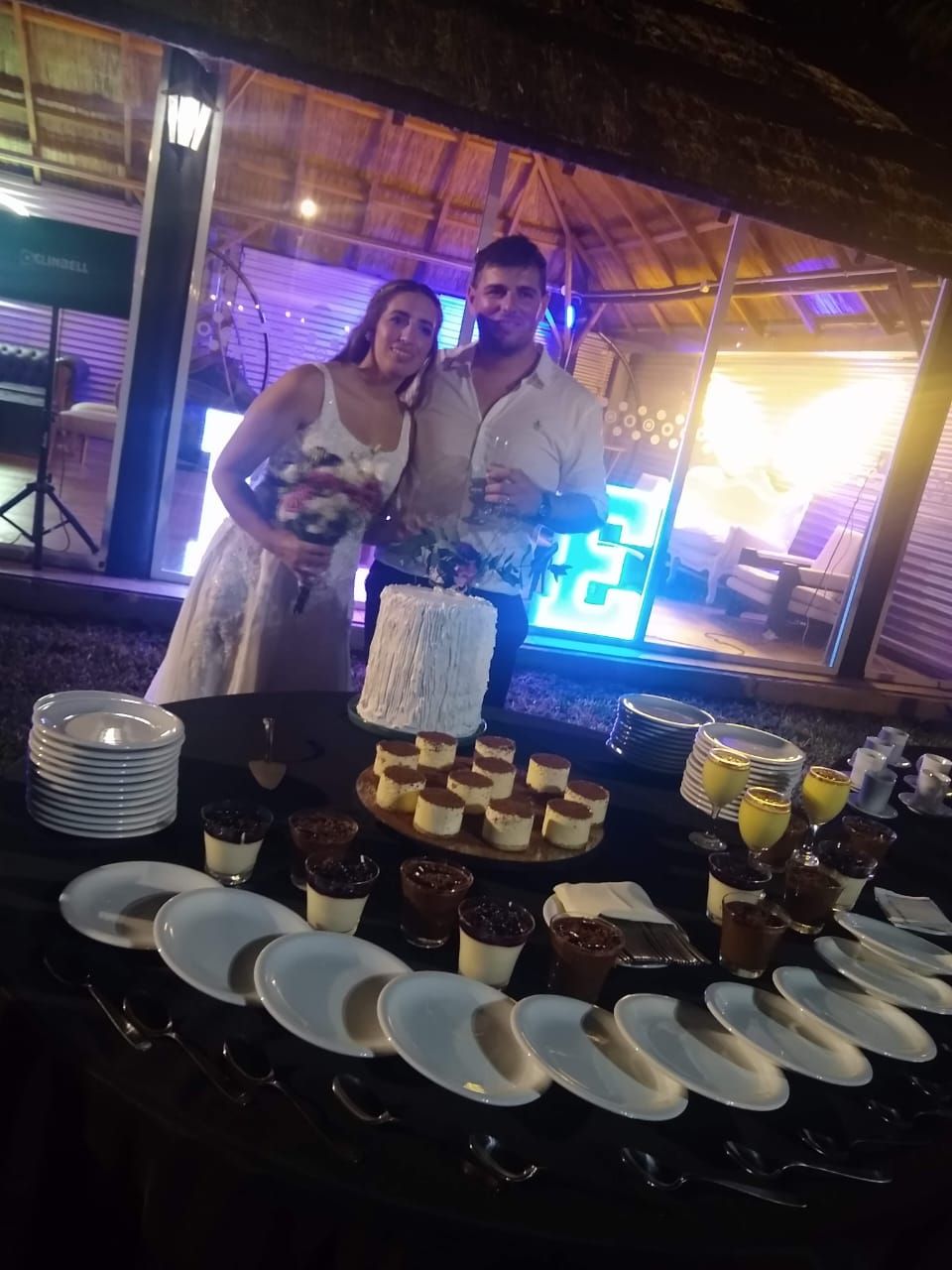 Una pareja de pie junto a una mesa de pasteles y postres en una recepción nocturna al aire libre con luces.