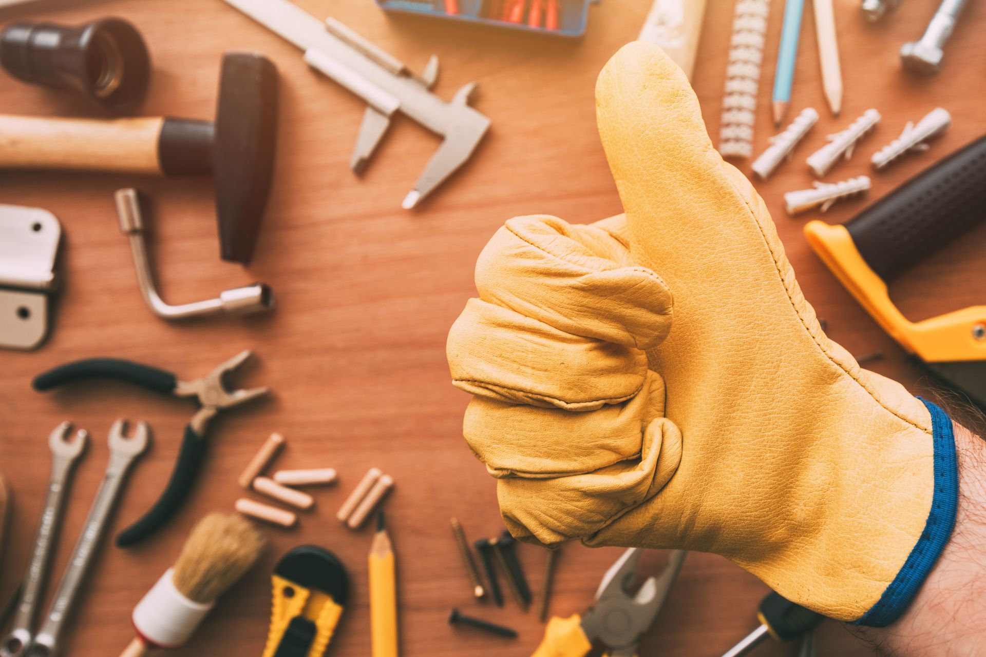 Hand in yellow work glove giving thumbs up; surrounded by tools on wooden surface.