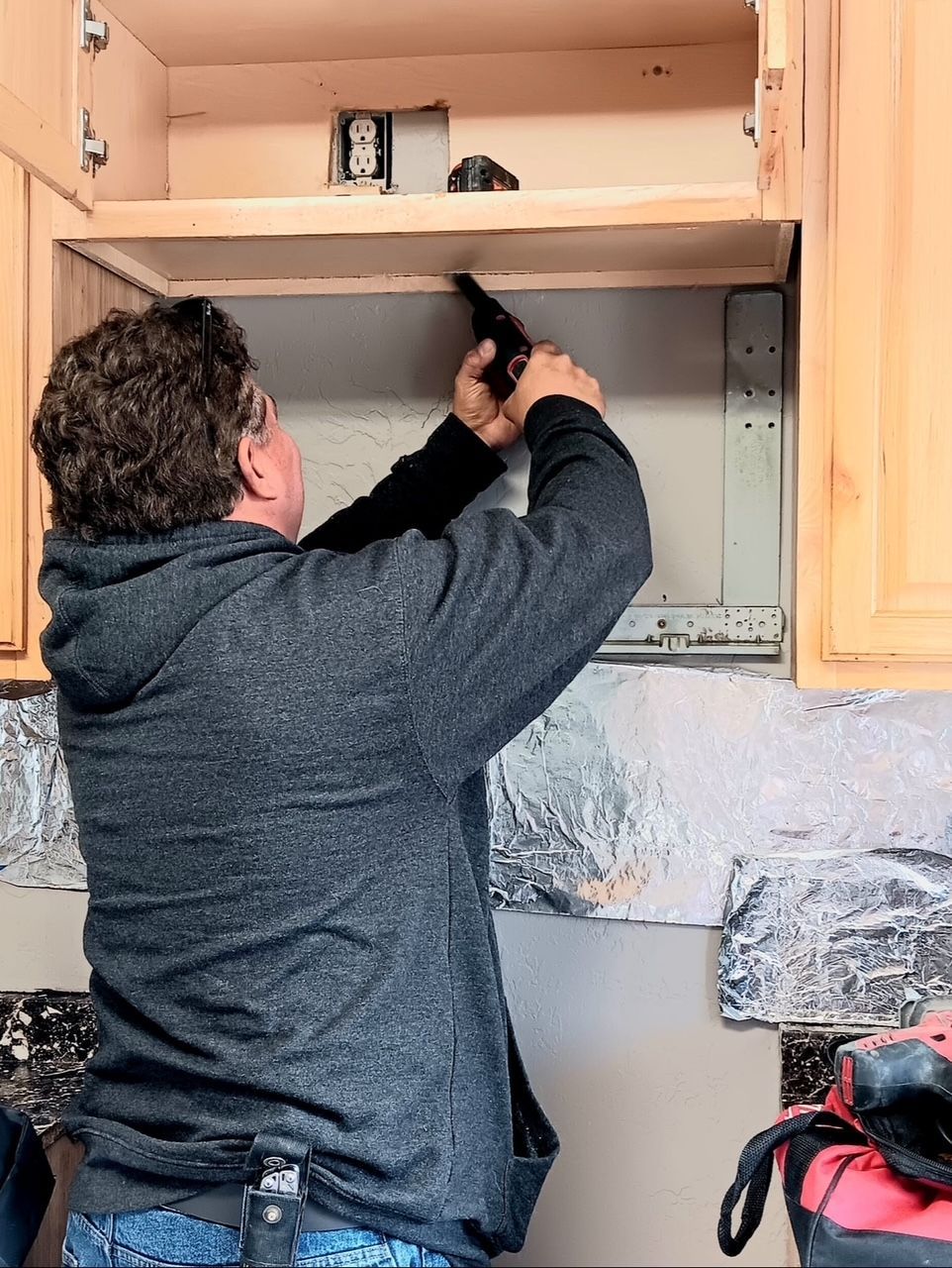 Person installing something inside a kitchen cabinet, wearing a gray hoodie, holding a tool.