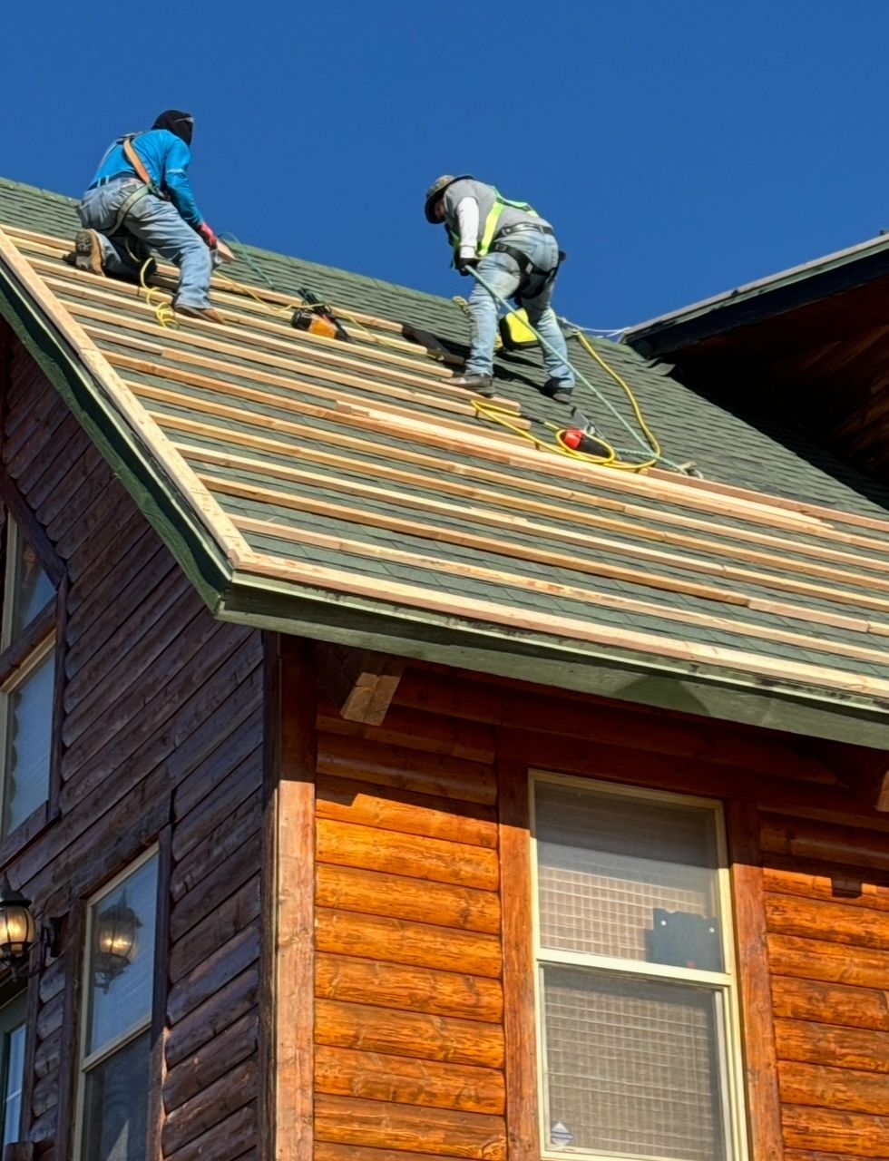 Two roofers on a roof installing new shingles; wood house, bright blue sky.