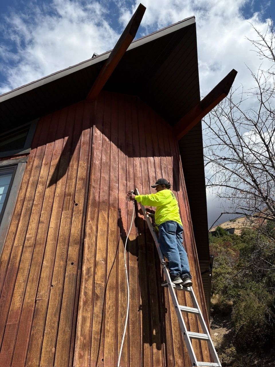 Person on ladder inspecting wood siding of a building with angled roof supports.