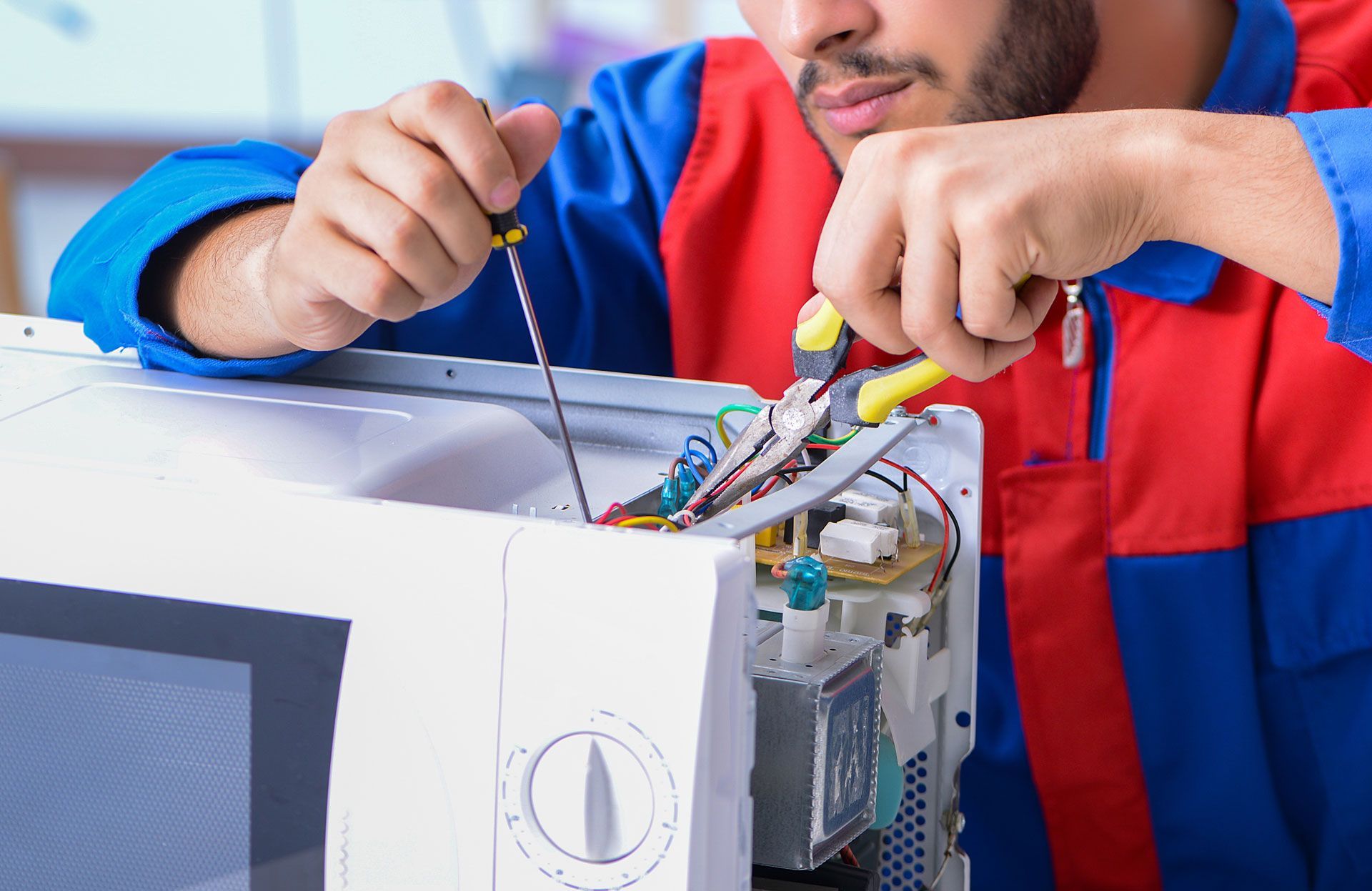 Man repairs a microwave with a screwdriver and pliers; blue and red work clothes.