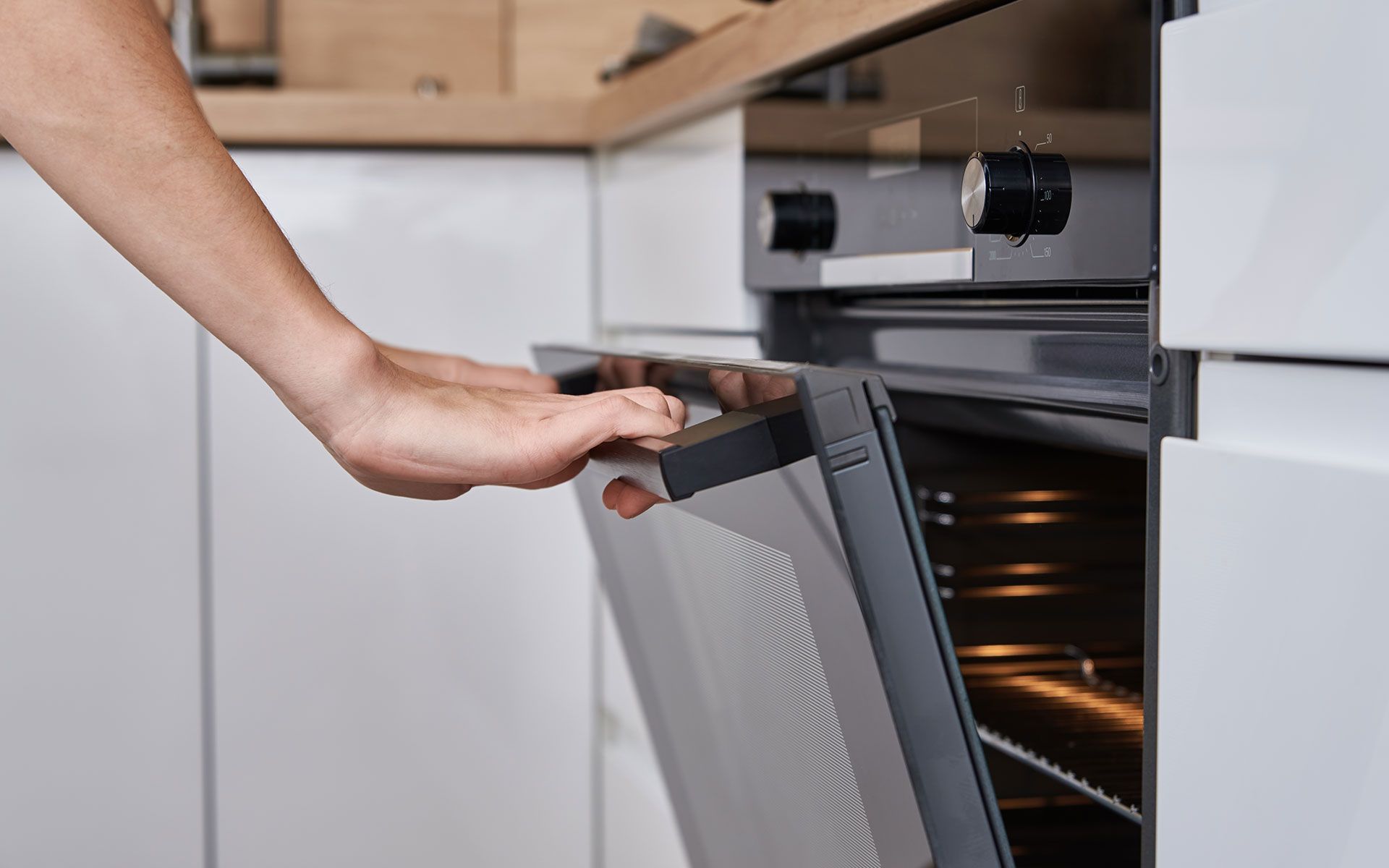 Hand opening an oven door in a white kitchen; the oven interior glows.