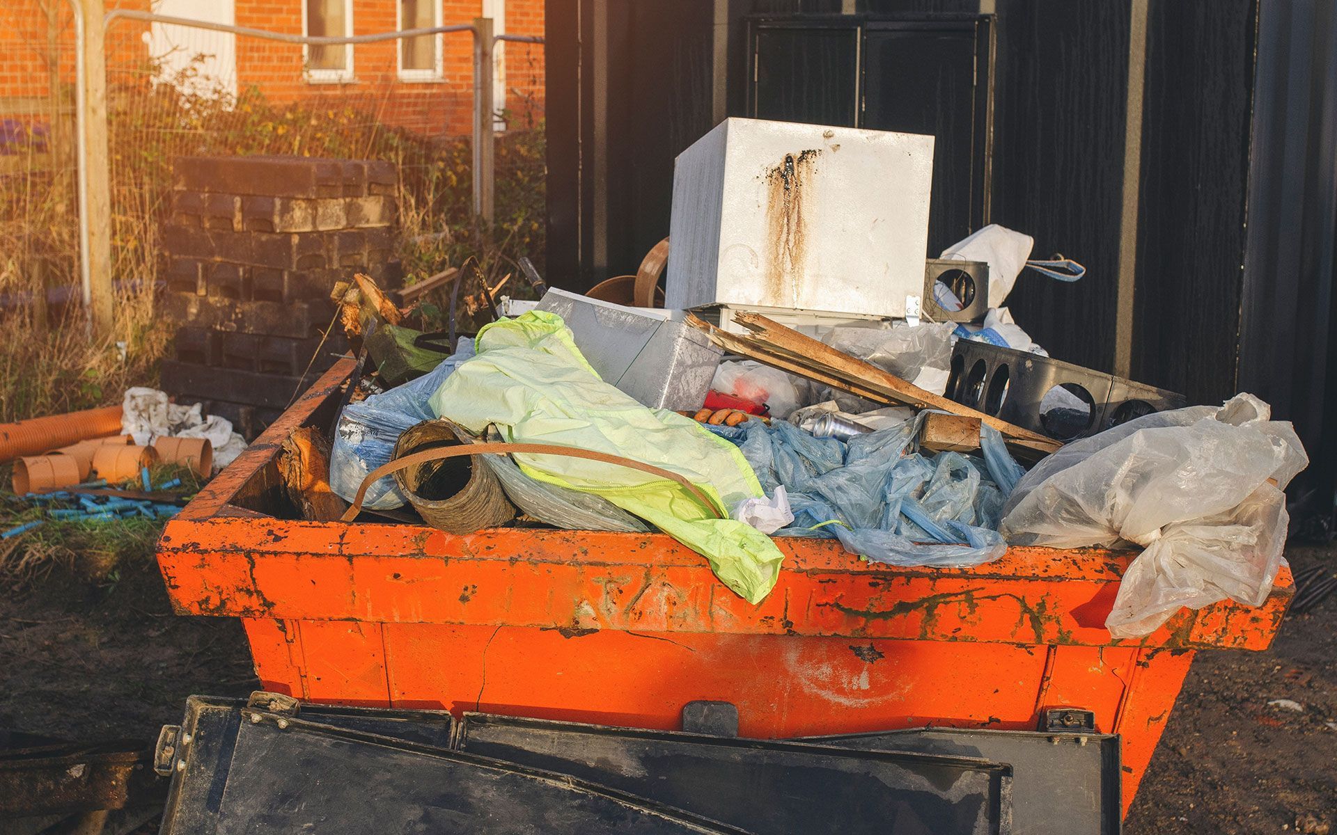 Orange dumpster overflowing with trash, near a fence and building.