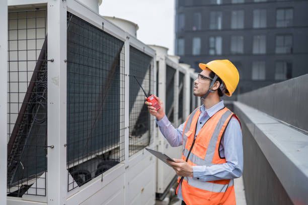 HVAC engineer in safety gear checks outdoor system with tablet and walkie-talkie.