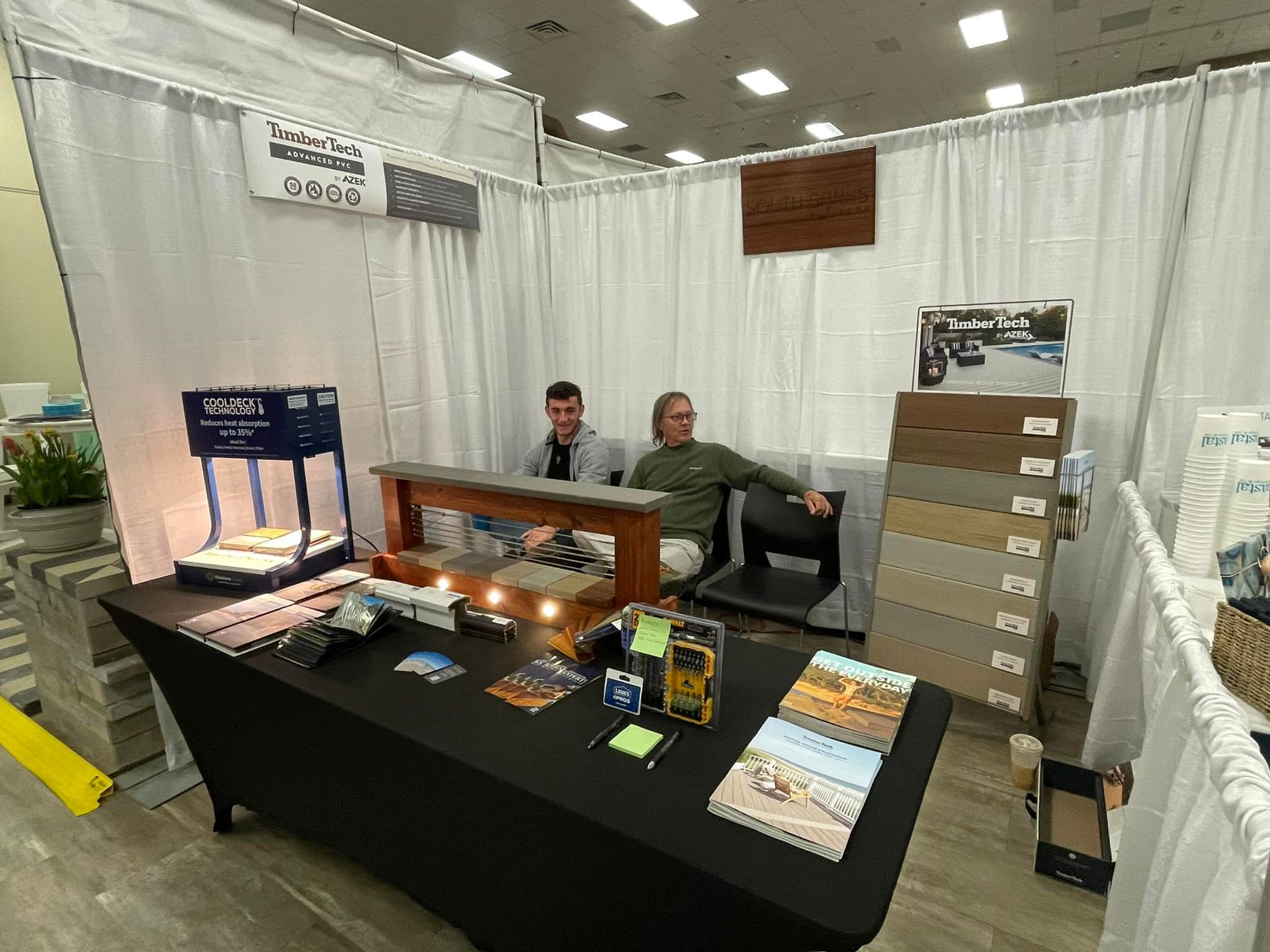 Two men are sitting at a table in a booth at a convention.