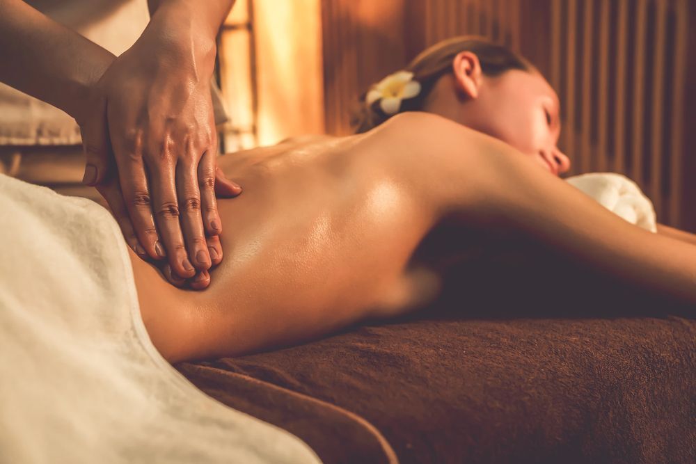 Persona tumbada boca abajo, con las manos entrelazadas, con una correa negra. Velas y plantas al fondo.