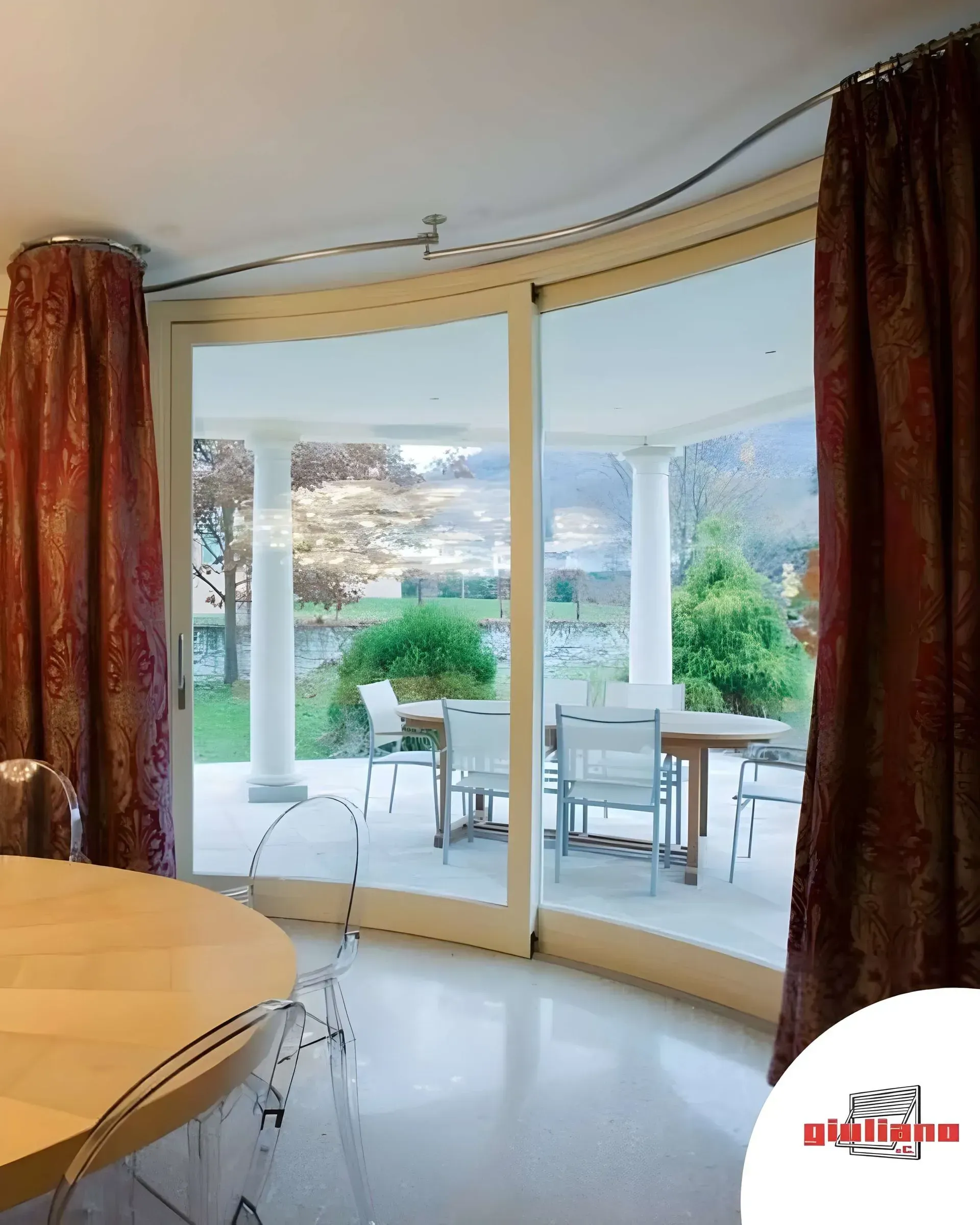 Le porte in vetro curvo si aprono su un patio. Le tende rosse incorniciano la vista dei posti a sedere all'aperto e del lago.