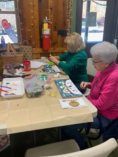Two older women are sitting at a table painting