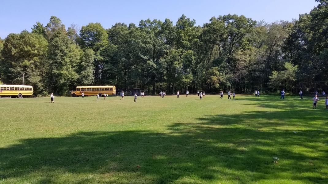 A group of people are playing soccer in a field with school buses in the background.