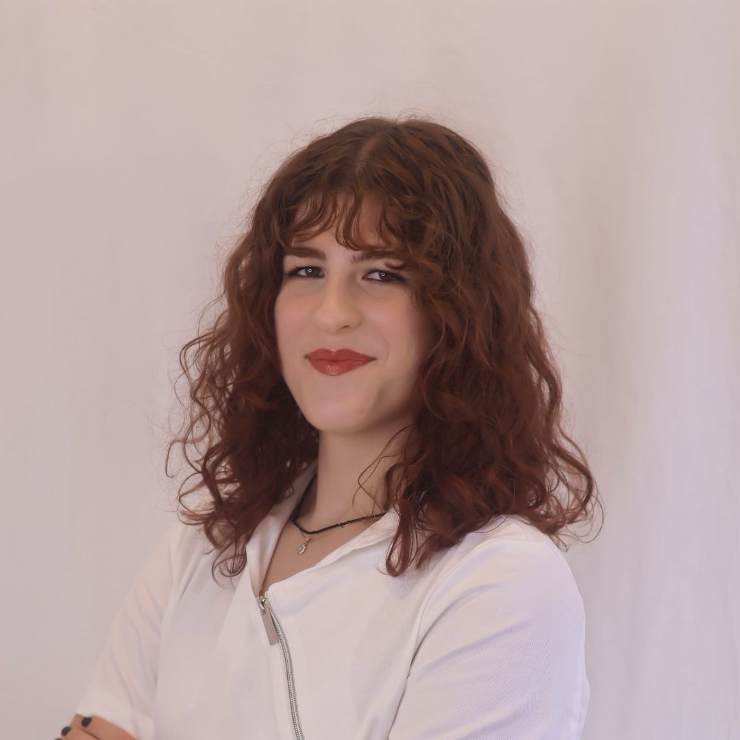 Woman with curly auburn hair, smiling, wearing white shirt, arms crossed, set against a white backdrop.