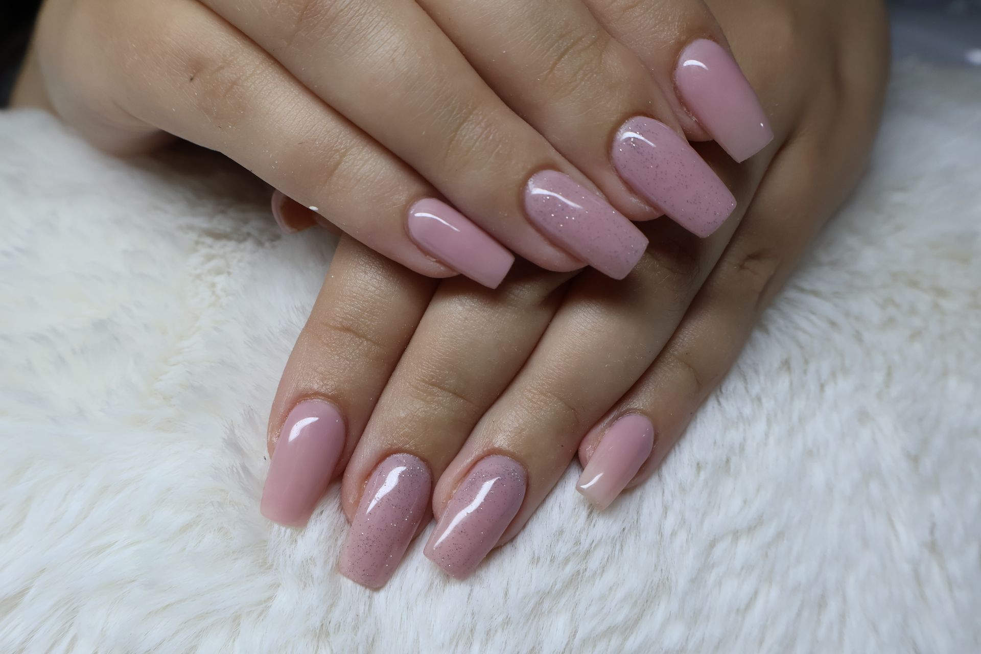 Hands with long, coffin-shaped, sparkly pink nails resting on white fuzzy fabric.