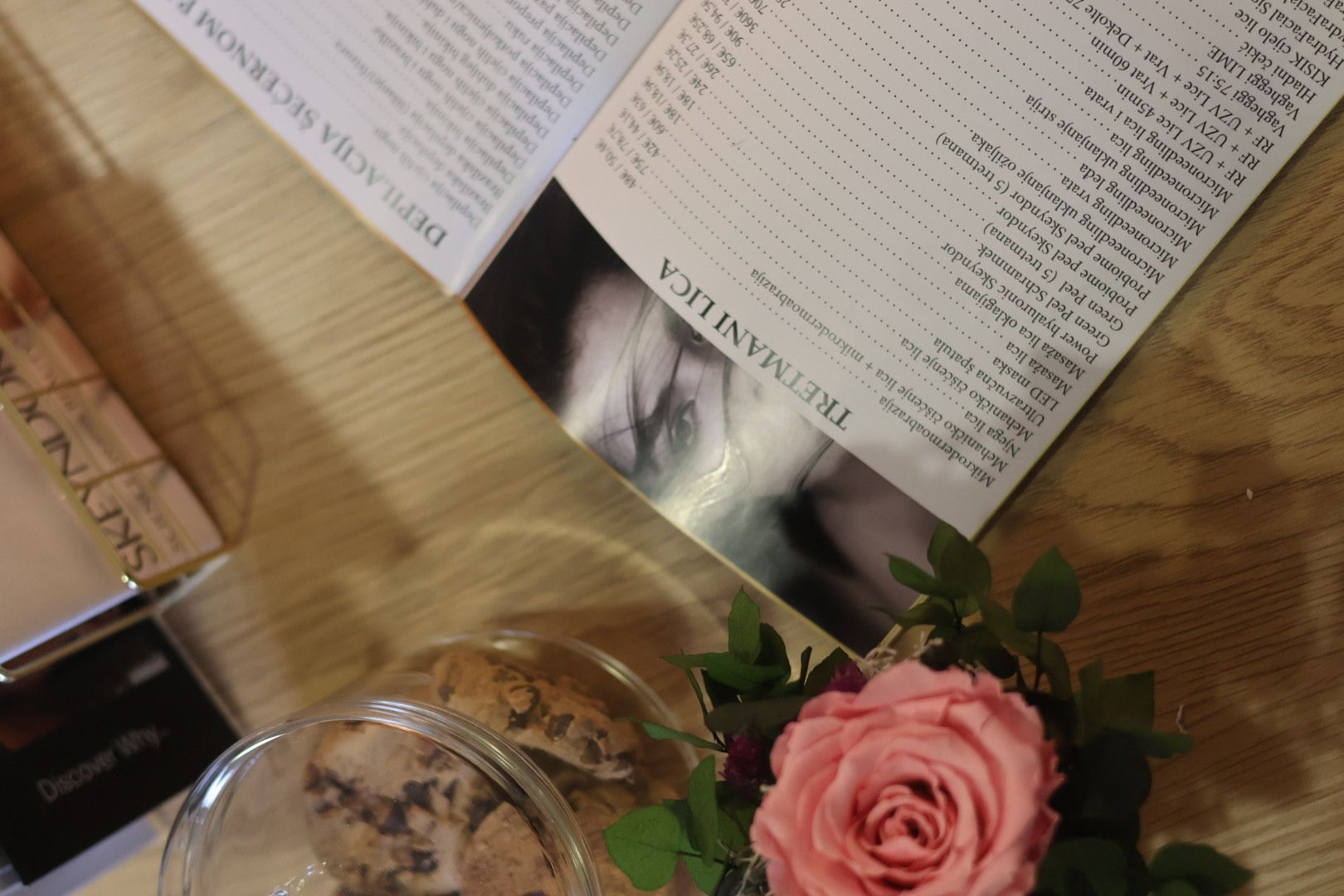 An open magazine, a pink rose, and beauty products are on a wooden surface.