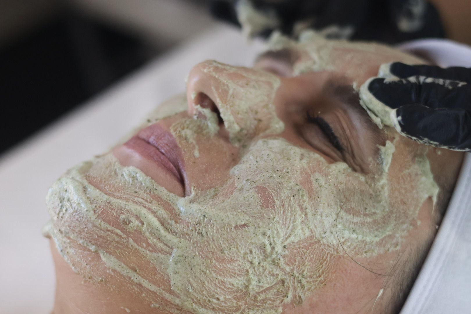 Person's face with a green facial mask applied in a spa setting; gloved hands are visible.