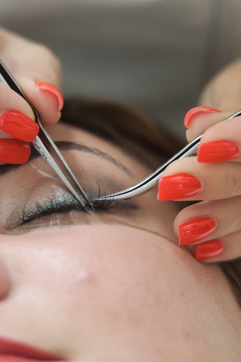A person receiving eyelash extensions. A technician uses tweezers to apply a lash. Red fingernails. LD BEAUTY CENTAR