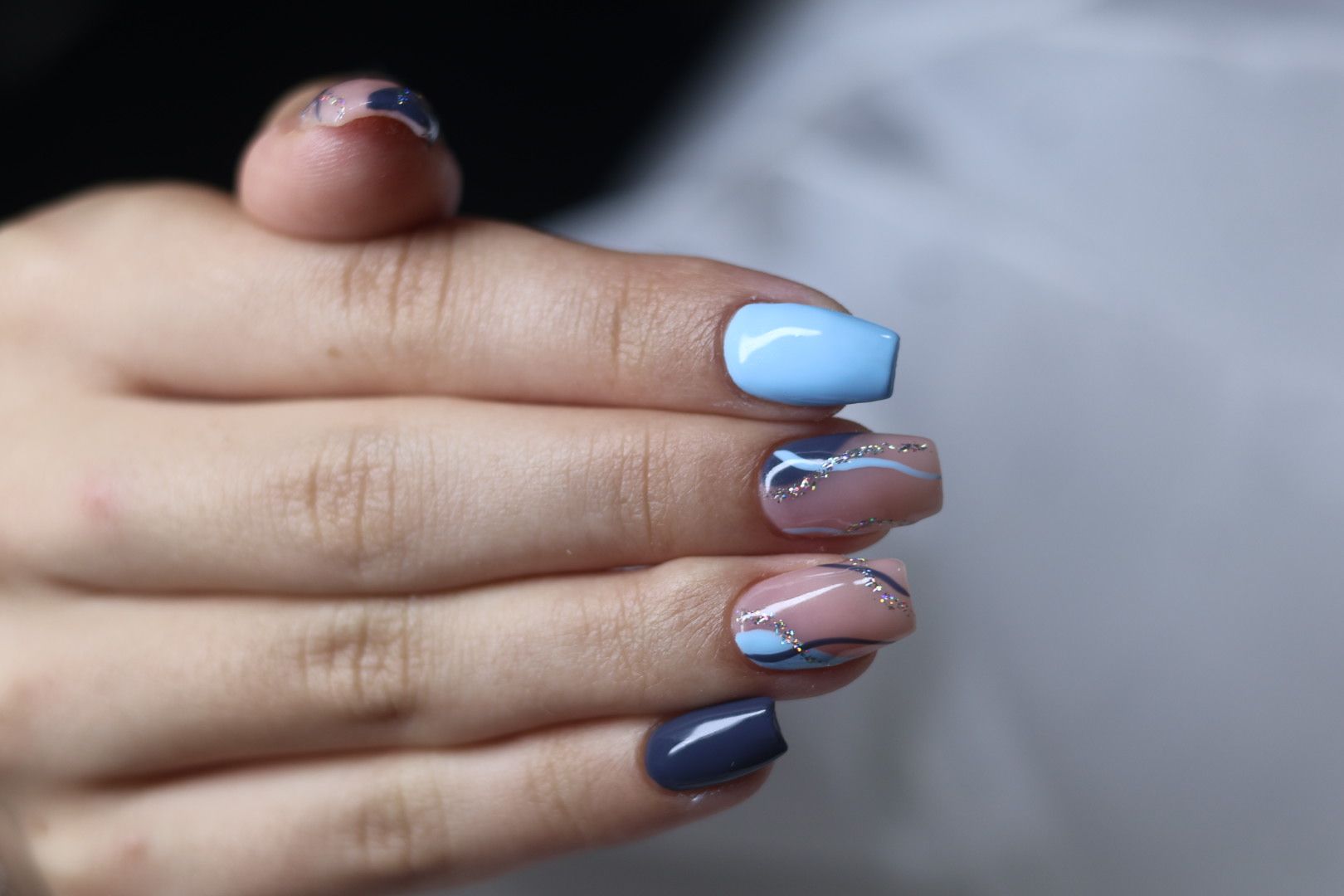 Hand with short, square nails painted in various blue and nude tones, with abstract designs. LD BEAUTY CENTAR