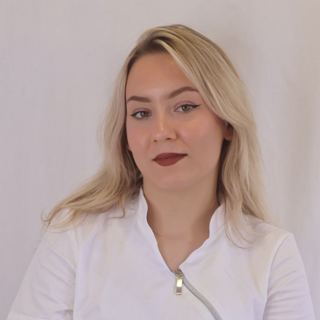 Blonde woman in white coat, dark lipstick, looking at the camera.