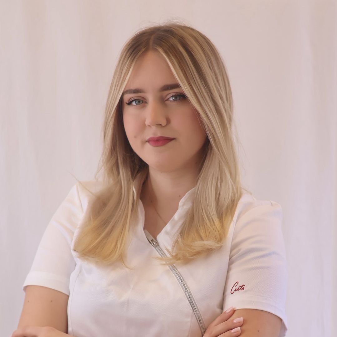 Woman with blonde hair, wearing a white uniform, arms crossed, looking at the camera.