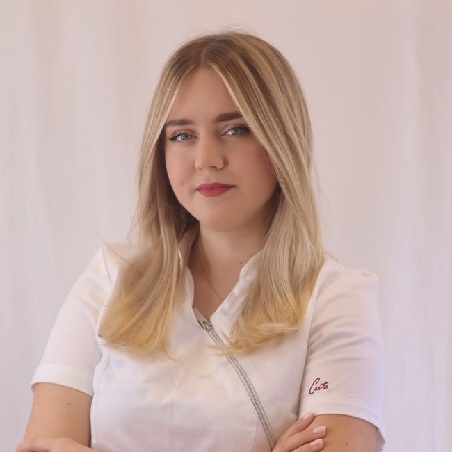 Woman with blonde hair, wearing a white uniform, arms crossed, looking at the camera.