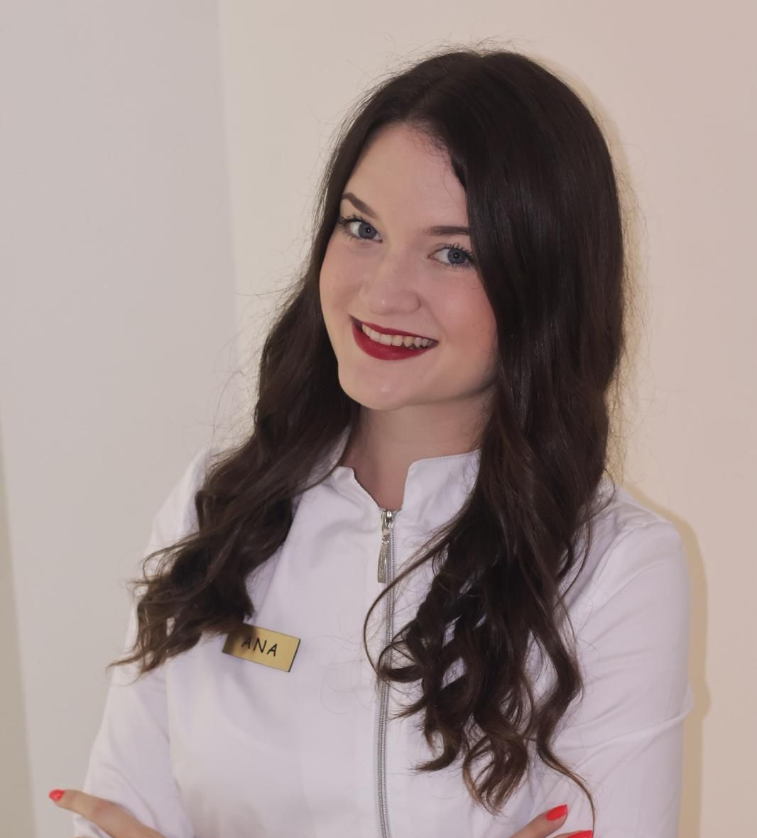 Woman in white uniform smiles, arms crossed; red lipstick, name tag 