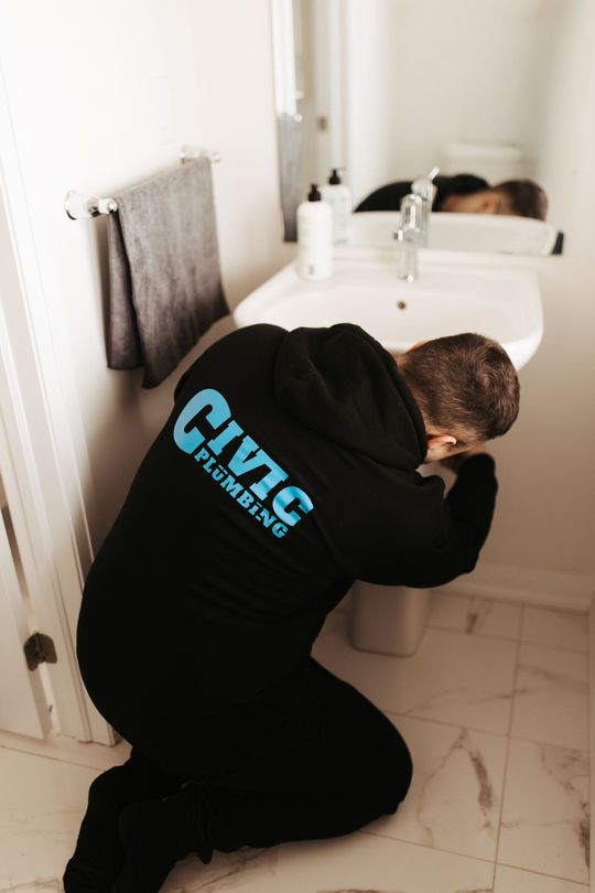 Plumber kneeling by a toilet, working on plumbing. Wearing a black sweatshirt with 
