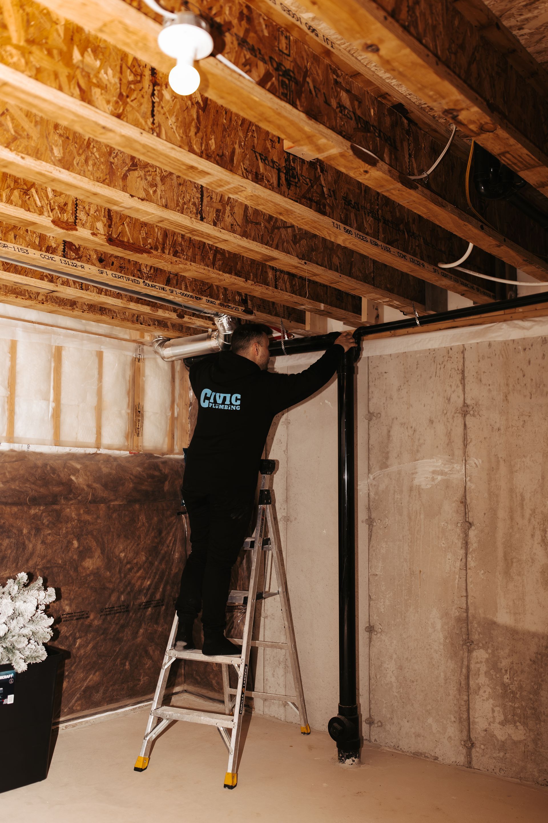 Person on a ladder installing black pipe in a basement. Interior shot with wooden beams and concrete wall.