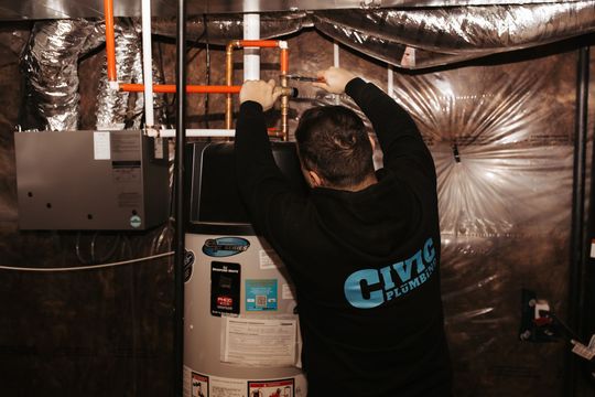 Plumber working on a water heater in a utility room; wearing a black shirt, using a wrench.