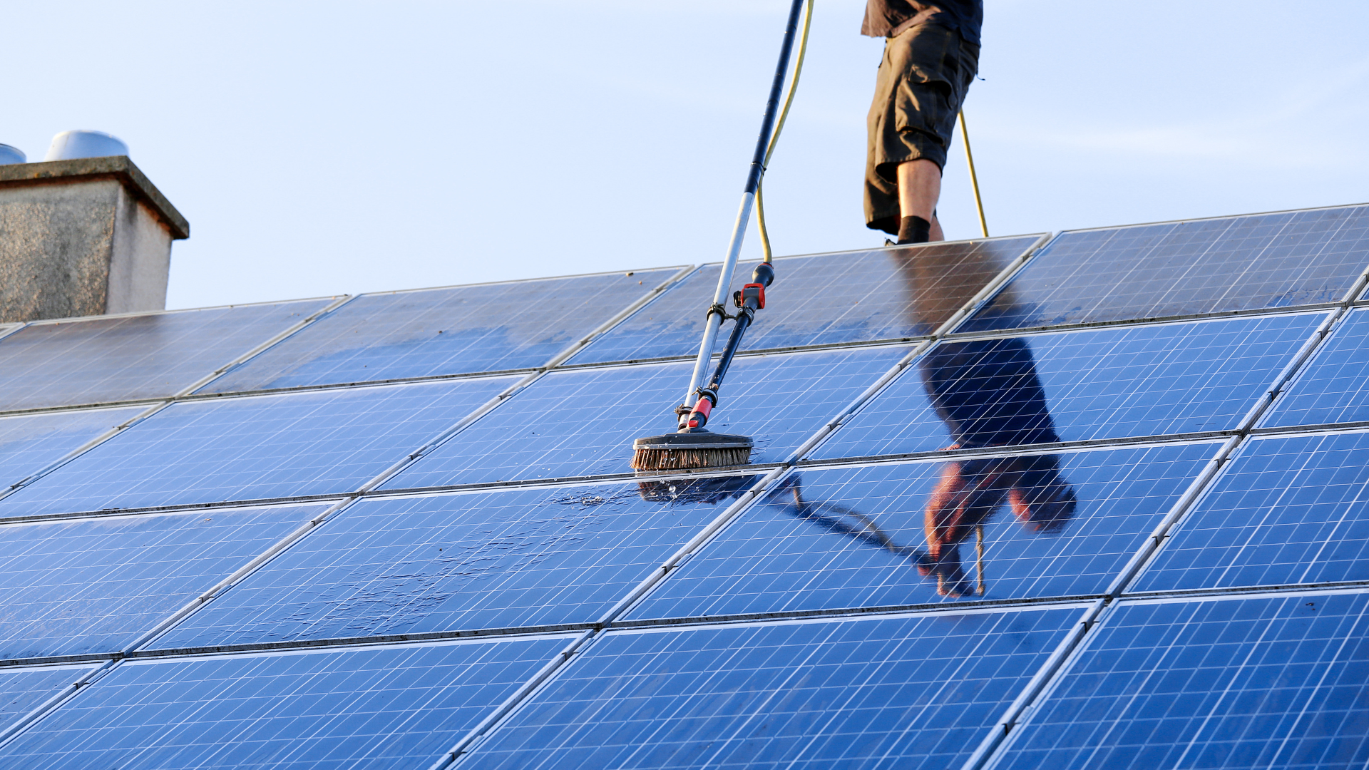 Person cleaning solar panels on a rooftop with a long-handled cleaning tool; sunny, blue sky.