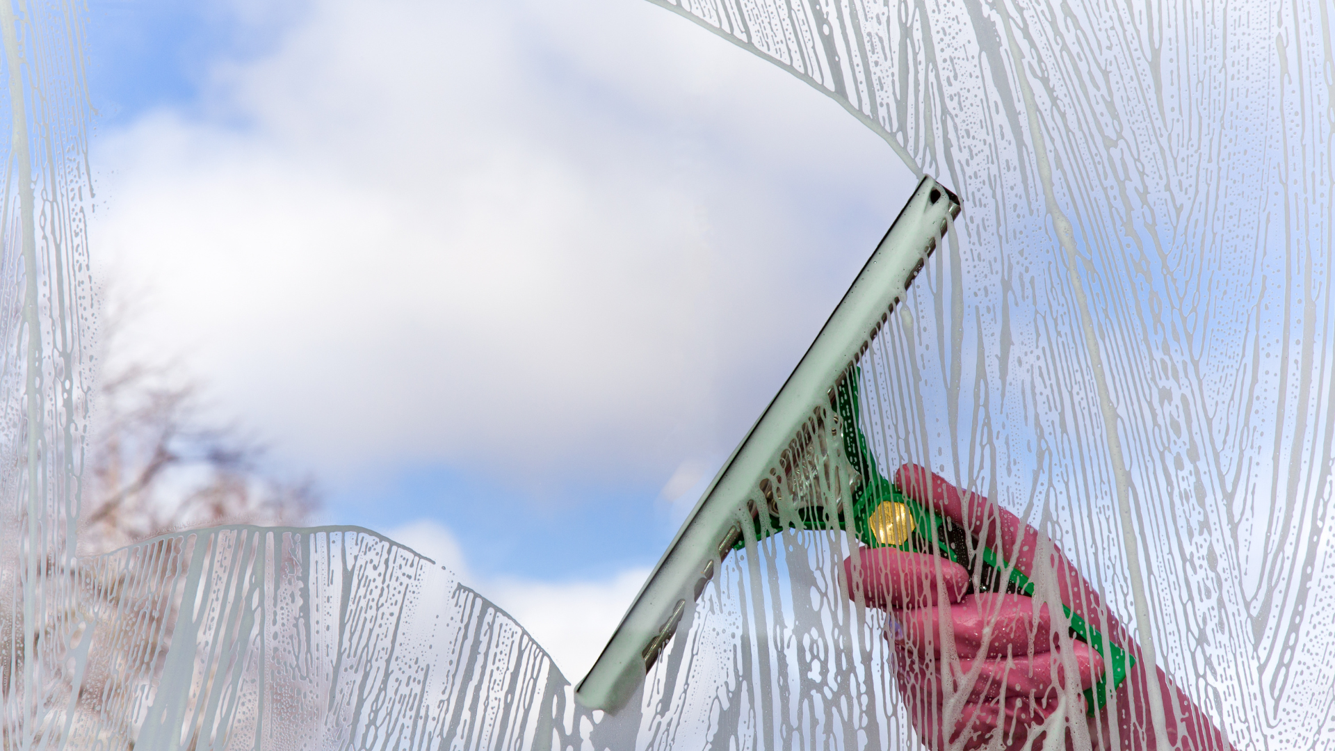 Hand wearing pink glove uses a squeegee to clean a window; sky visible through cleared area.