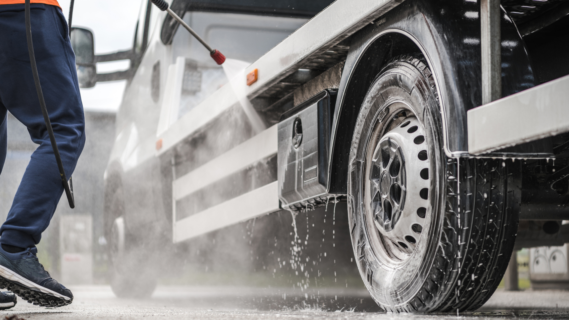 Person pressure washing a white delivery truck's tire and side on a wet, concrete surface.