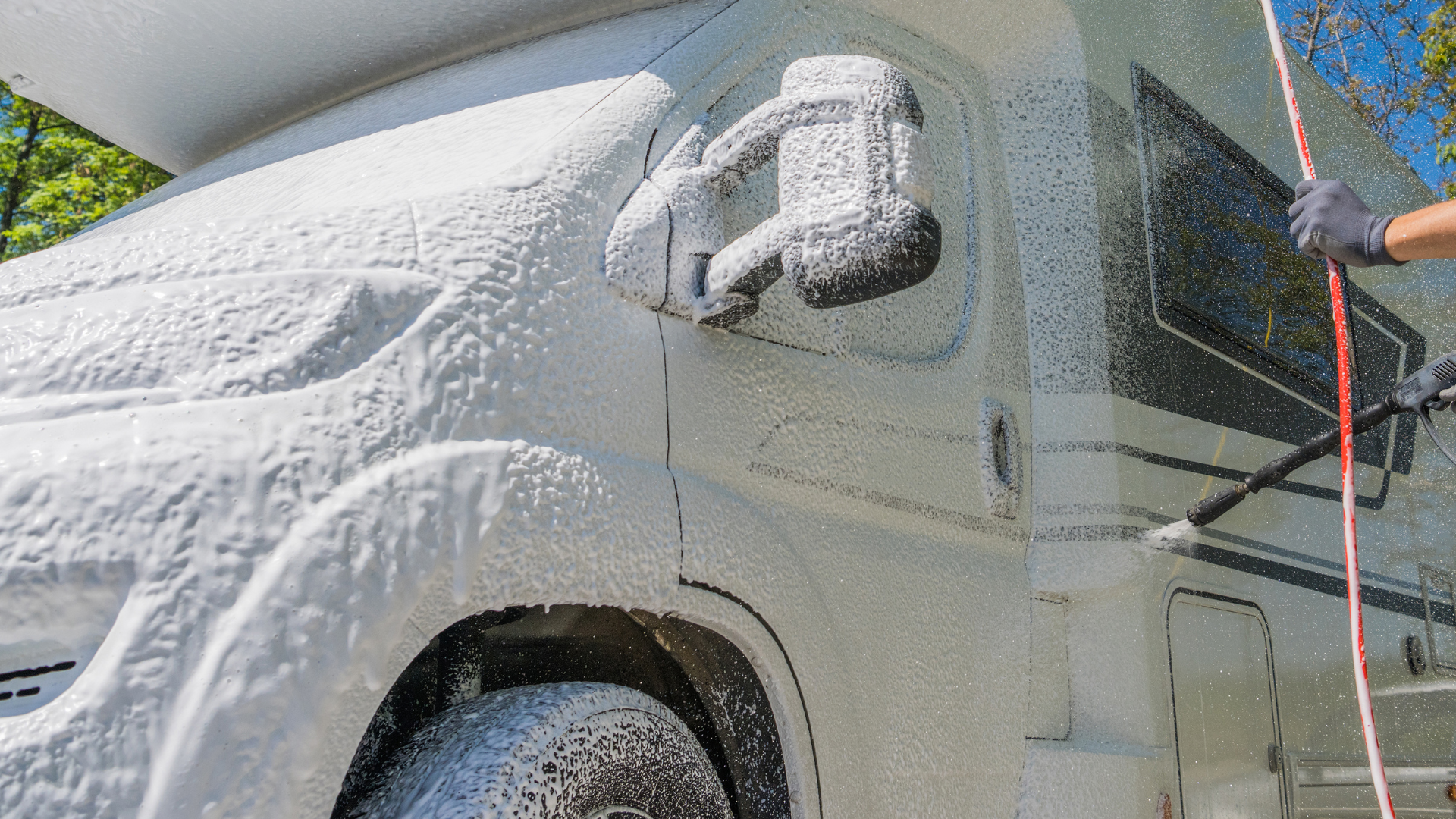 A white RV being covered in white foam, likely for cleaning. A gloved hand holds a spray wand.