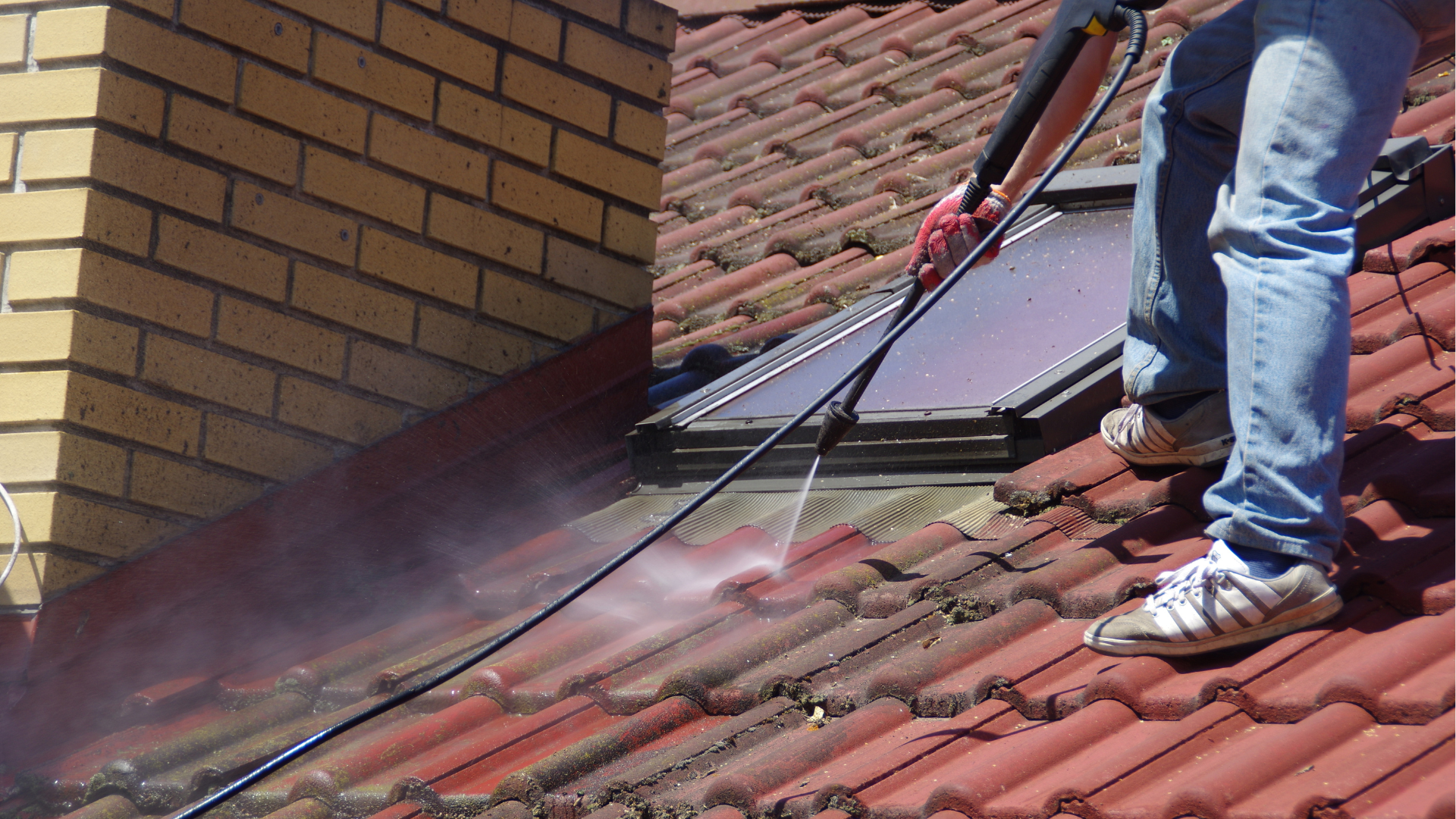 Man pressure washing a red tiled roof near a chimney and skylight.