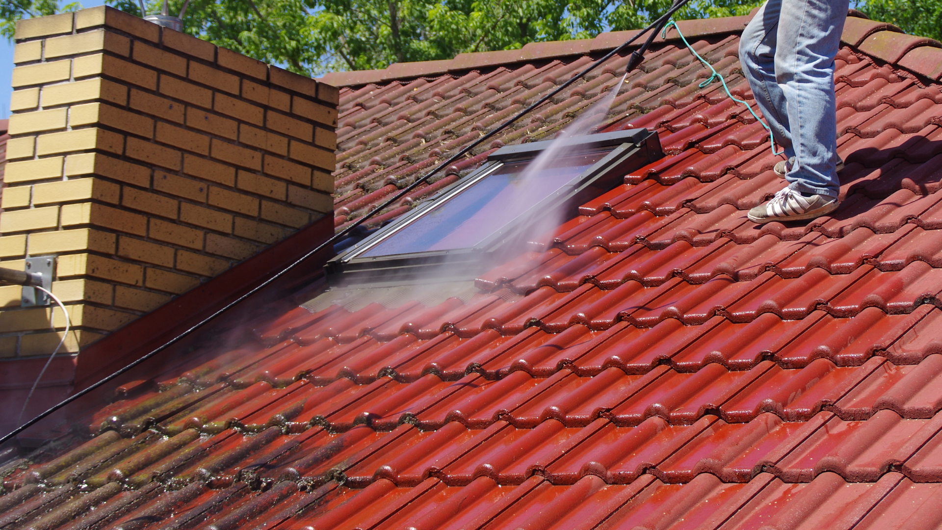 Person pressure washing a red tiled roof with a skylight, chimney in the background.