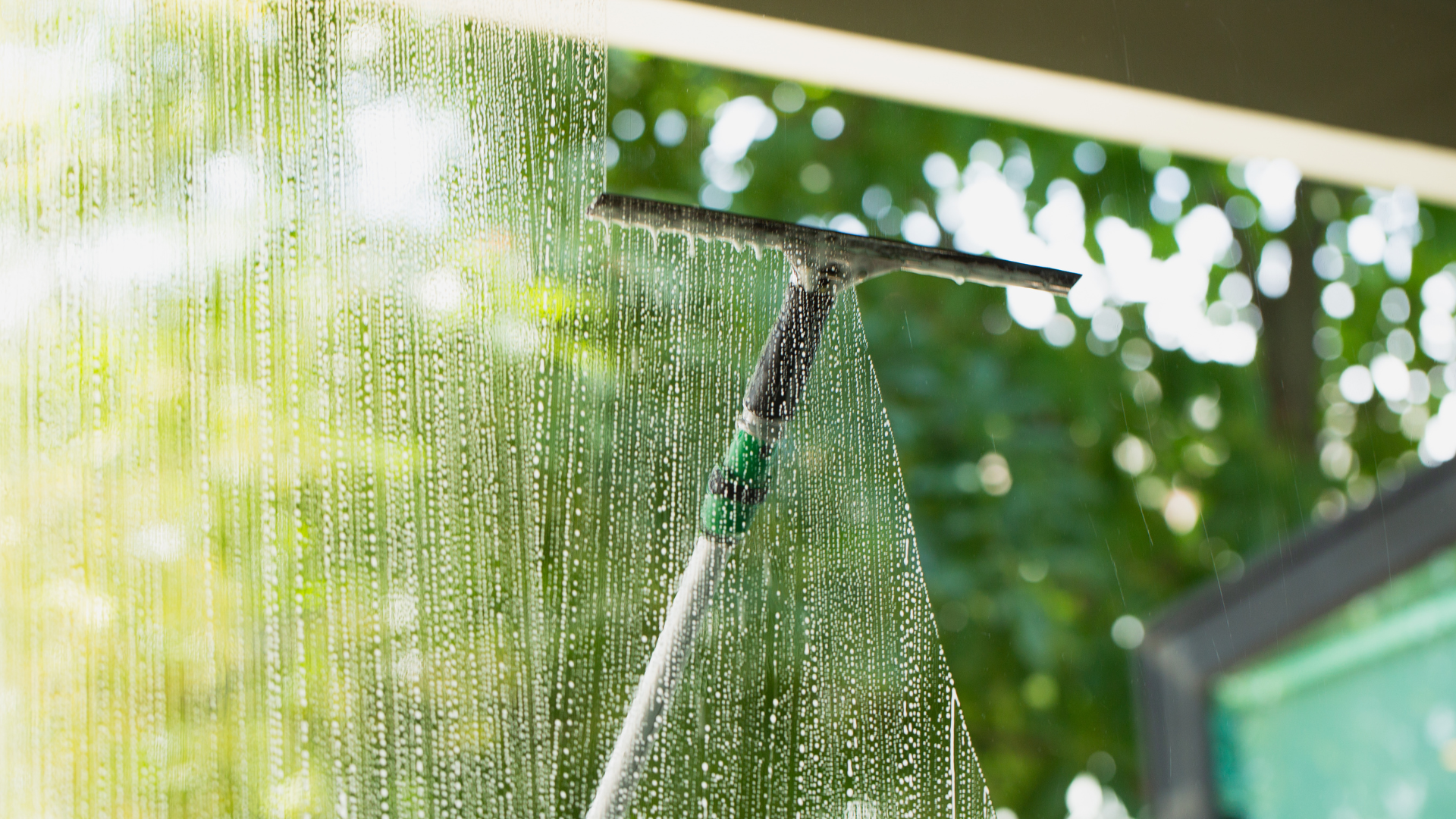 Squeegee cleaning a wet window, streaks of water visible, greenery blurred in background.