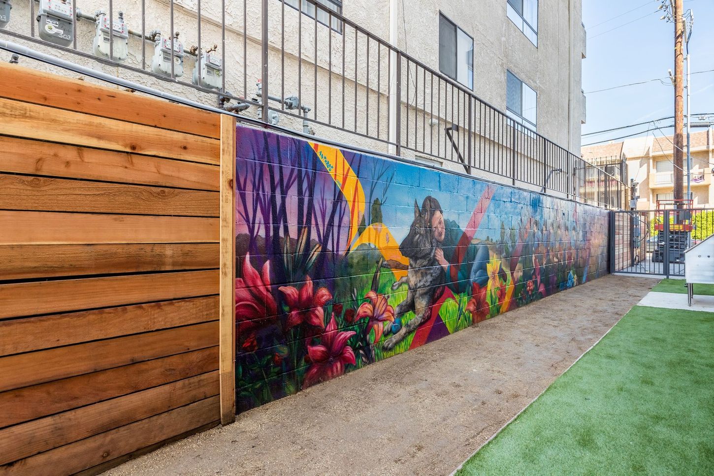 A vibrant mural on a concrete wall with flowers, trees, and a person holding a sword. Wooden fence on the left.