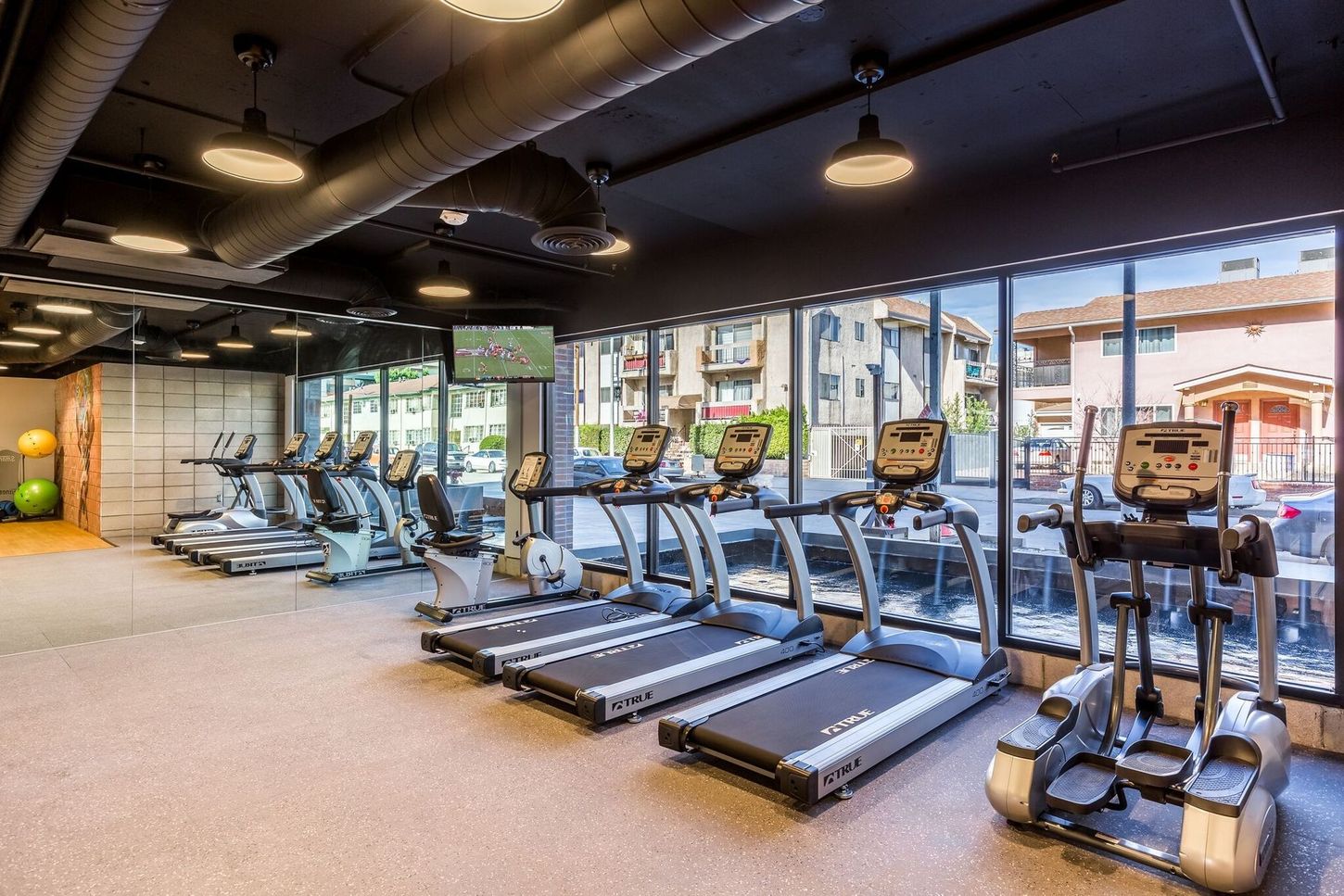 Gym with treadmills, elliptical, exercise bikes, and a TV; large windows offer an outdoor view.
